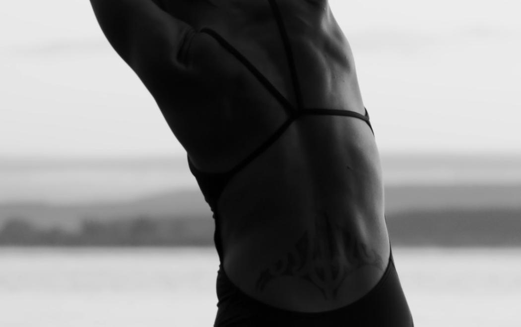 The back of a women swimmer, wearing black togs and stretching to the left.